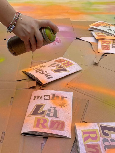 Das Bild zeigt in Nahaufnahme eine Hand, die mit einer Sprühdose Farbe auf Kartons und Zines sprüht.