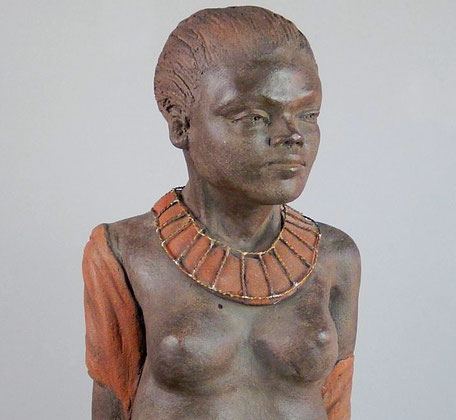 Le buste dénudé d'un femme africaine les cheveux plaqués en arrière avec son regard apaisé et sa parure en métal autour du cou.