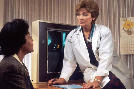 Abbildung einer Ärztin aus dem Jahr 1990 zeigt eine Ärztin die auf dem Tisch gegenüber einer Patientin sitzt.