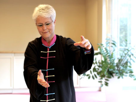 Trainerin zeigt eine Armhaltung des Qigong. Sie trägt einen pinken Qigonganzug. Beide Arme werden auf Kopfhöhe vor dem Körper gehalten. Die Hände halten einen imaginären Qiball. Die Trainerin schaut zu Ihnen mit freundlichem, entspannten Lächeln.