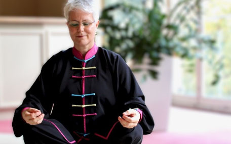 Meditationslehrerin sitzt im Schneidersitz in einem schwarzen Qigonganzug mit farbigen Applikationen auf dem Boden. Die Hände liegen auf den Knien, die Finger sind zur Meditationsgeste geschlossen. Die Augen sind geschlossen, ein Lächeln im Gesicht. 02