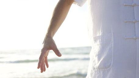 Repräsentativ für das Qigong-Einzeltraining ist der Ausschnitt eines Mannes gezeigt, der an einem Strand mit dem Meer im Hintergrund eine stehende Position einnimmt. Er trägt einen weißen Qigonganzug, man sieht einen Teil des Rumpfes und einen Arm.