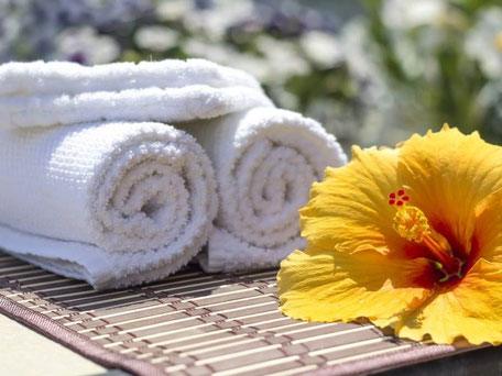 Sie sehen eine gelbe Hibiskusblüte auf einem Bambusset. Links der Blüte liegen zwei weiße Handtuchrollen. Im unscharfen Hintergrund Blüten. Hibiskus und Handtücher sind Symbole für das Angebot Chinesische Energiemassage.