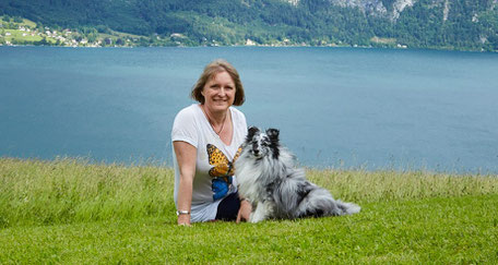 Astrid Huber mit kleinem weiß-schwarz-grauem Langhaarhund in Wiese sitzend. Dahinter Attersee.
