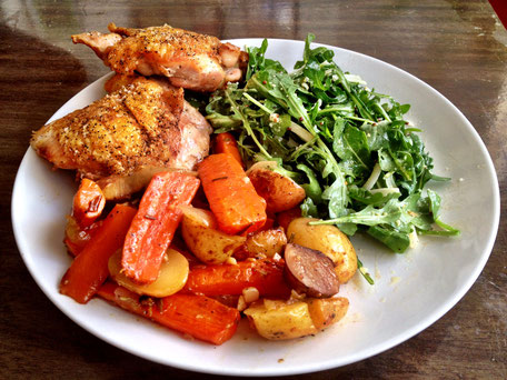 A plate of grilled chicken thigh, roasted carrots and potatoes and a side of rocket salad on a white porcelain plate set against a dark wooden table