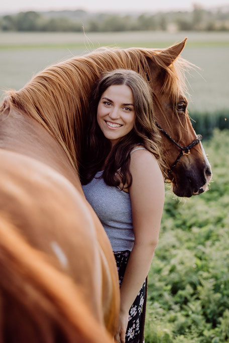 Junge Frau mit braunen Haaren steht neben ihrem braunen Pferd, das seinen Kopf über ihre Schulter legt und zur Seite schaut
