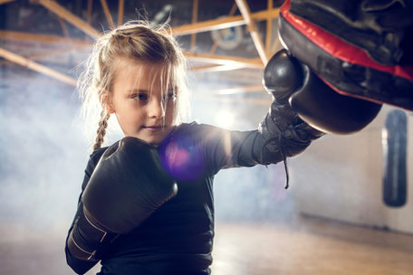 Ein 6 Jahriges Mädchen in Kickbox Uniform, schlägt mit Boxhandschuhe auf eine Pratze. Zu sehen ist auch, das sich das Mädchen Konzentriert und Spaß bei der Übung hat. 