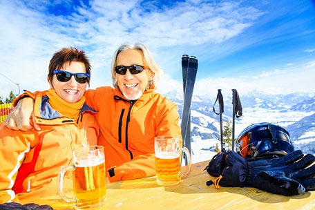 Pärchen mit Bier auf der Piste beim Wintersport in den Bergen, Skifahren mit der passenden Reiseversicherung