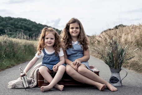 Zwei Mädchen sitzen fröhlich auf einem Vintage-Koffer inmitten der Natur in Mülheim. Ein unbeschwerter Moment voller Abenteuerlust und Kindheitszauber beim Outdoor-Kindershooting.