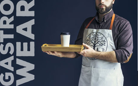 Mann mit Bart trägt Schürze und hält ein Tablett mit Kaffeebecher, neben ihm steht „GASTRO WEAR“.