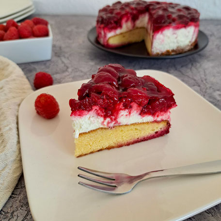 Ein Stück Himbeertraum auf einem weißen Teller, im Hintergrund die angeschittene Torte und eine Schale mit frischen Himbeeren