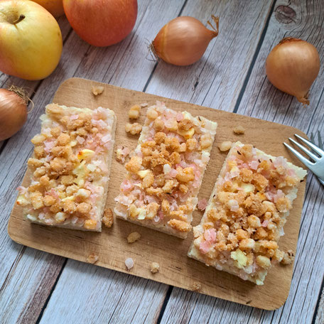 Drei Stück Speckkuchen auf einem Holzbrett, darum herum liegen Äfpel und Zwiebeln