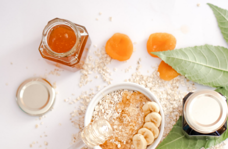 Vogelperspektive von Müslischale mit Müsli und Bananen, daneben getrocknete Marillen und ein offenes Glas Honig
