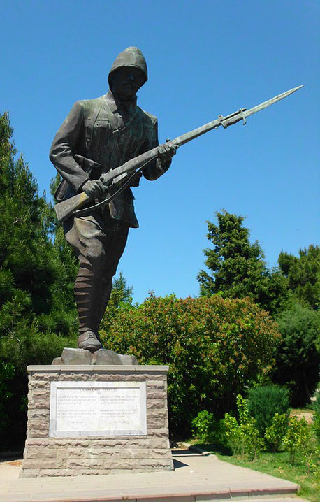 A bronze statue of a World War I soldier holding a rifle with bayonet stands on a stone pedestal in a landscaped park.
