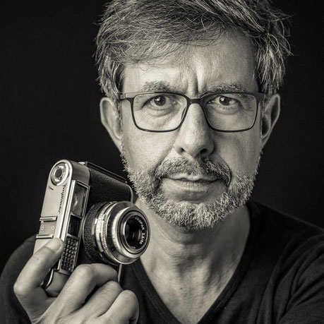 Un portrait en noir et blanc du photographe Philippe Colin, il nous regarde dans les yeux en tenant dans sa main droite un appareil photo ancien