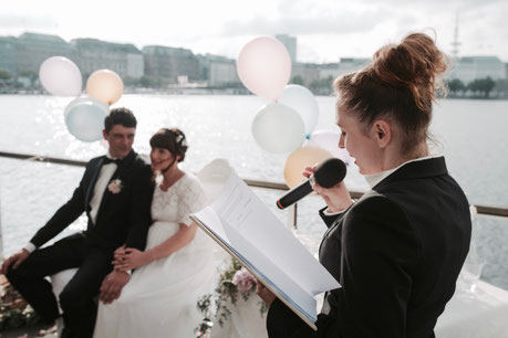 Constanze Marienfeld spricht bei einer Trauung auf der Alster ins Mikro – im Vordergrund das verschwommene Brautpaar, im Hintergrund glitzert das Wasser