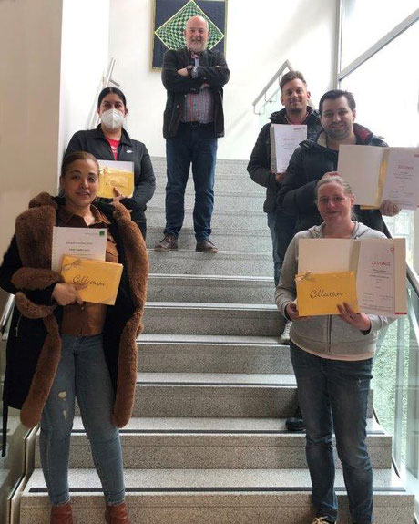 Gruppe von Teilnehmer:innen mit Lehrabschlusszeugnissen steht auf einer Treppe und hält stolz ihre Urkunden in der Hand