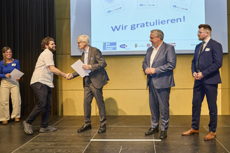 Absolvent nimmt Urkunde entgegen – Gratulanten im Hintergrund