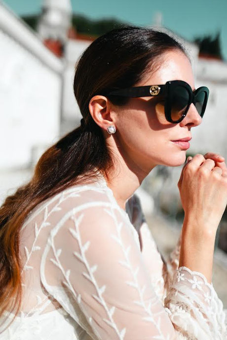 Frau in weißer, bestickter Bluse und großer schwarzer Sonnenbrille im Sonnenlicht. Sie trägt silberne Perlenohrringe, der Fokus liegt auf ihrem eleganten Profil.