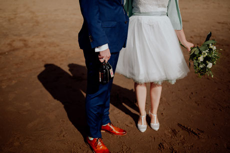 Füsse stehen am Elbstrand, man sieht den Anzug und das Brautkleid und eine Hand mit zwei Flaschen Astra. Freie Trauung Hamburg, Constanze Marienfeld