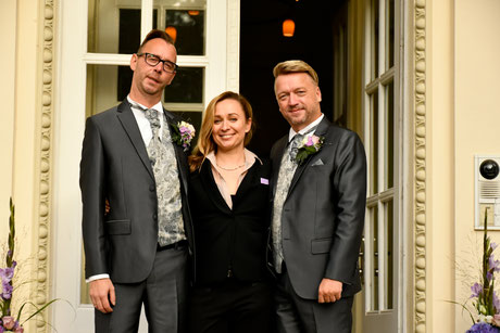 Constanze Marienfeld steht entspannt, nach der Trauung, zwischen ihren beiden Bräutigamen vor einer großen Flügeltür die ins Schloß führt, alle drei lächeln.