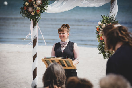Traurednerin Constanze Marienfeld seht vor ihm Traubogen uns strahlt in die Gastreihen, im Hintergrund rauscht die Ostsee