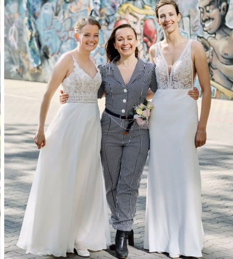 Constanze Marienfeld lächelt stolz, im Arm hält sie ihr Brautpaar, zwei Frauen, beide im Brautkleid