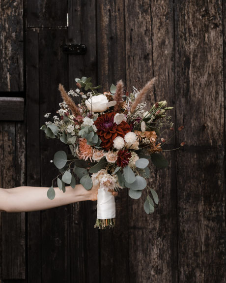 Bouquet verschiedener Blumen, ein Herbstblumenstrauss, er wird von einer Hand vor eine von Sonne & Regen ergrauten Stalltür gehalten