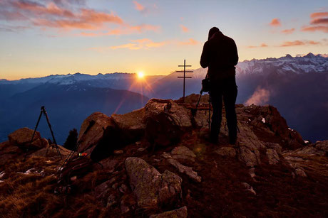 Ein Fotograf mit Stativ steht im Gegenlicht eines Sonnenuntergangs auf einem Gipfel in den herbstlichen Alpen, im Hintergrund sind verschneite Berggipfel und ein Gipfelkreuz zu sehen