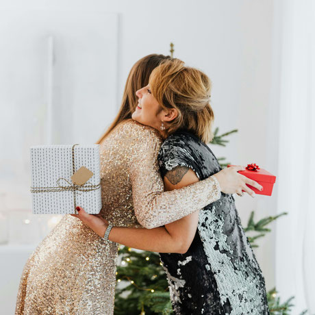 Zwei Frauen in festlicher Kleidung umarmen sich vor dem Weihnachtsbaum und tauschen Geschenke – Symbol für Freude und Verbundenheit.