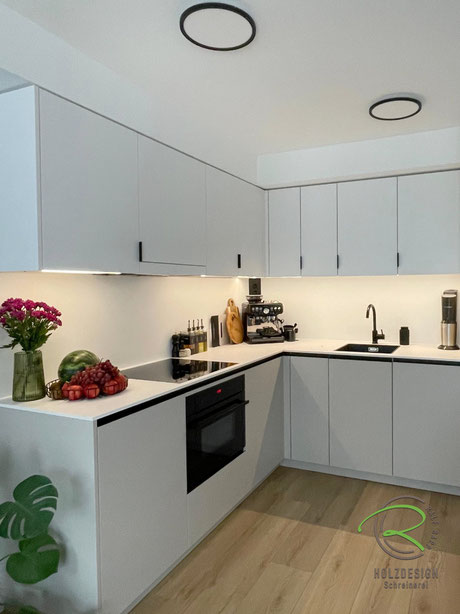 Helle, kleine Wohnküche über Eck mit schwarzen Designelementen wie Griffleisten, Armatur & Spüle als besonderes Designelement, mit deckenhohen Kücheneinbauschränken, im Oberschrank integrierte Dunstabzugshaube u. filigrane Kompaktarbeitsplatte in weiß 