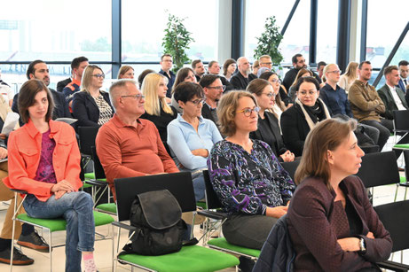 Engagiertes Publikum in einem modernen Konferenzraum mit Glasfassade. Männer und Frauen unterschiedlichen Alters folgen dem Vortrag.