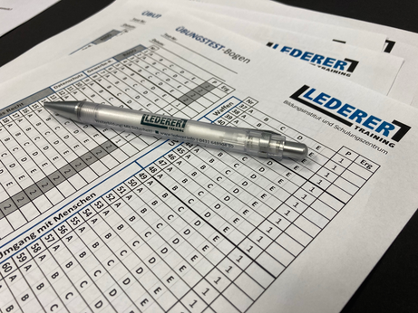 Übungstest-Bögen für die IHK-Sachkunde-Prüfung nach § 34a GewO (im Volksmund: "34a-Schein"). Auf dem Tisch liegen mehrere Übungsbögen übereinander. Auf den Bögen und auf einem darüber liegenden Kugelschreiber steht das Logo von LEDERER_training.  