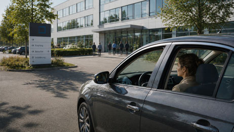 Eine Frau sitzt auf einem Firmenparkplatz noch im Auto, während andere bereits im Gebäude sind. Das Bild steht für den Moment zwischen Ankunft und dem nächsten Schritt.