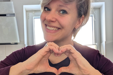 Rebecca Paschen lächelt in die Kamera, deutet mit den Händen vor ihrem Oberkörper ein Herz an. Sie steht im Bad, hat kurze blonde Haare und ein lilafarbenes Oberteil an. 