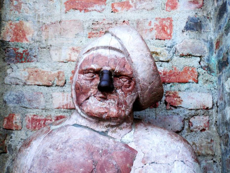 Nahaufnahme der historischen Büste des „Steinernen Mannes“ (Stoinarne Ma) in Augsburg vor einer alten Ziegelmauer. Das stark verwitterte Gesicht aus rötlichem Stein zeigt eine charakteristische Beschädigung an der Nase und eine schiefe Kopfbedeckung. Ein 