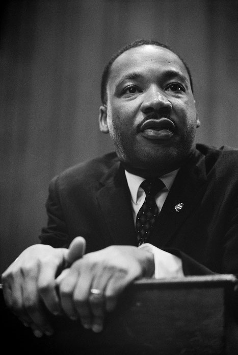 Black-and-white close-up of a man in a suit and tie, leaning forward with his hands resting on a surface, looking thoughtfully ahead.