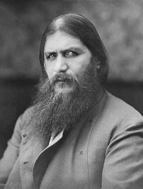 A man with intense eyes, long hair, and a full beard stares directly at the camera, wearing a buttoned coat in this grayscale portrait.