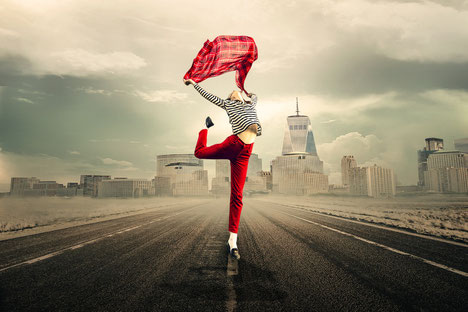 Sur une route goudronnée, une personne debout sur une jambe s'étirant vers le ciel en tenant une chemise colorée rouge entre ses bras tendus, sur fond de grande ville avec ses immeubles et une tour pointant sa flèche vers le ciel