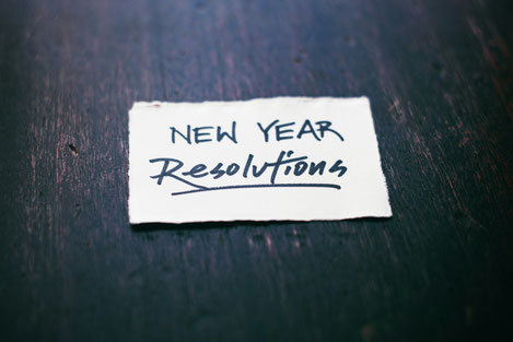 Auf einem Zettel, dre auf einer Holztischplatte liegt, steht handgeschrieben: New Year Resolutions