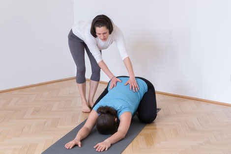 Schwangere Frau in Childs Pose / Balasana mit Yogalehrerin die mit ihren sanft aufgelegten Händen die tiefe Bauchatmung unterstützt und anleitet