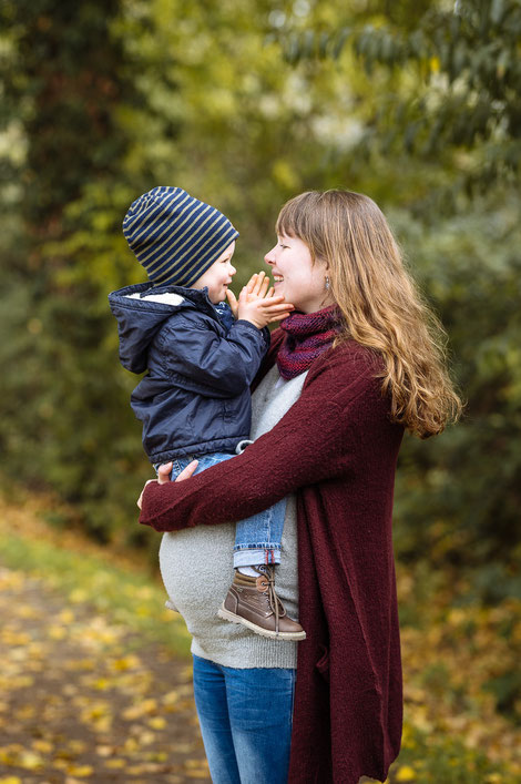 Schwangere Frau trägt lächelnd ihren kleinen Sohn auf dem Babybauch im herbstlichen Umfeld.