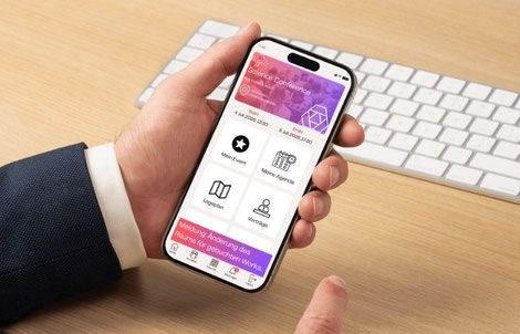 Eine Hand hält ein Smartphone, auf dem eine Übersichtsseite einer Event-App zu sehen ist. Im Hintergrund ist eine Tastatur auf einem Tisch zu sehen.