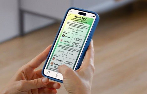 Hände halten ein Smartphone. Auf dem Display ist eine Agenda von einer Veranstaltung, mit verschiedenen Programmpunkten, zu sehen.