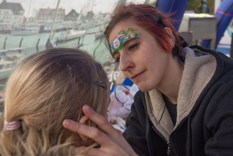 Pearl sitzt vor einem Kind und schminkt ihm ein buntes Motiv. Dabei hat sie einen aufgemalten Osterhasen auf der Stirn.