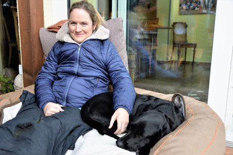 Frau im Rollstuhl mit Winterjacke Nina Hoffmann draußen auf der Terrasse mit schwarzer schlafender Labrador Hündin