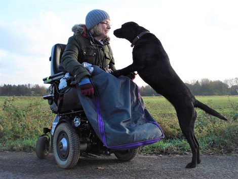 Frau Nina Hoffmann im Rollstuhl mit Mütze und Brille mit schwarzer Labrador Hündin auf der Wiese im Hintergrund Wald