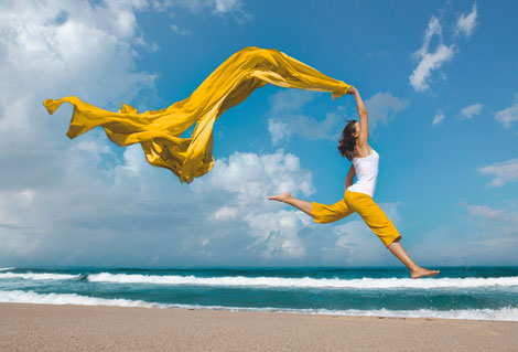 Eine Frau in senfgelbener Hose und mit senfgelbenem Schal springt am Strand vor Freude in die Höhe