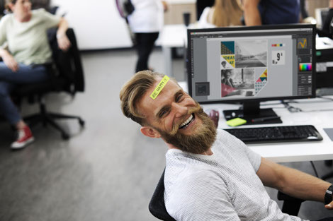 Homme à son bureau de travail, souriant et joyeux avec un post-it sur le front avec la mention "soit heureux"