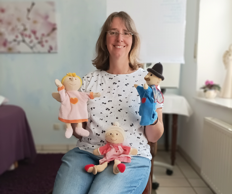 Geistheilerin Ina Paulus sitzt auf einem Stuhl und hält Handpuppen in den Händen